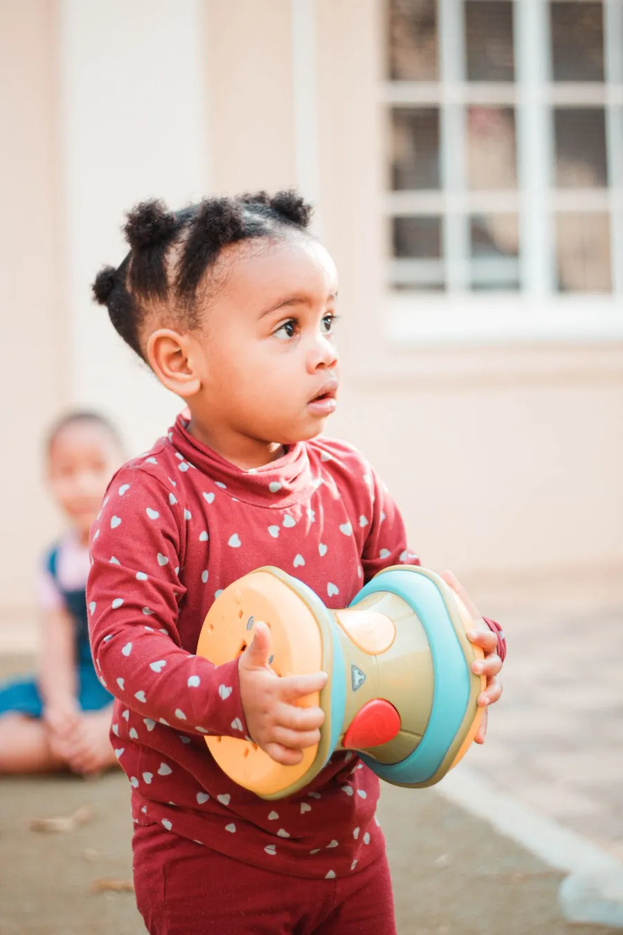 A toddler in a red heart-print top holding a colourful toy outdoors