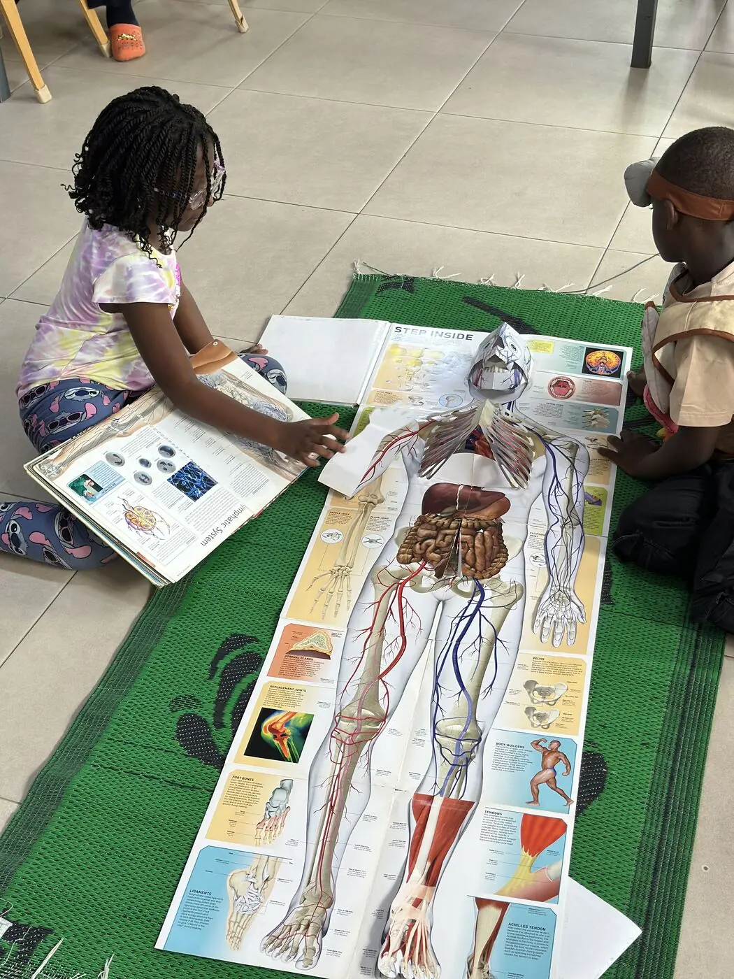 Two children exploring a life-size human body chart laid out on a floor mat